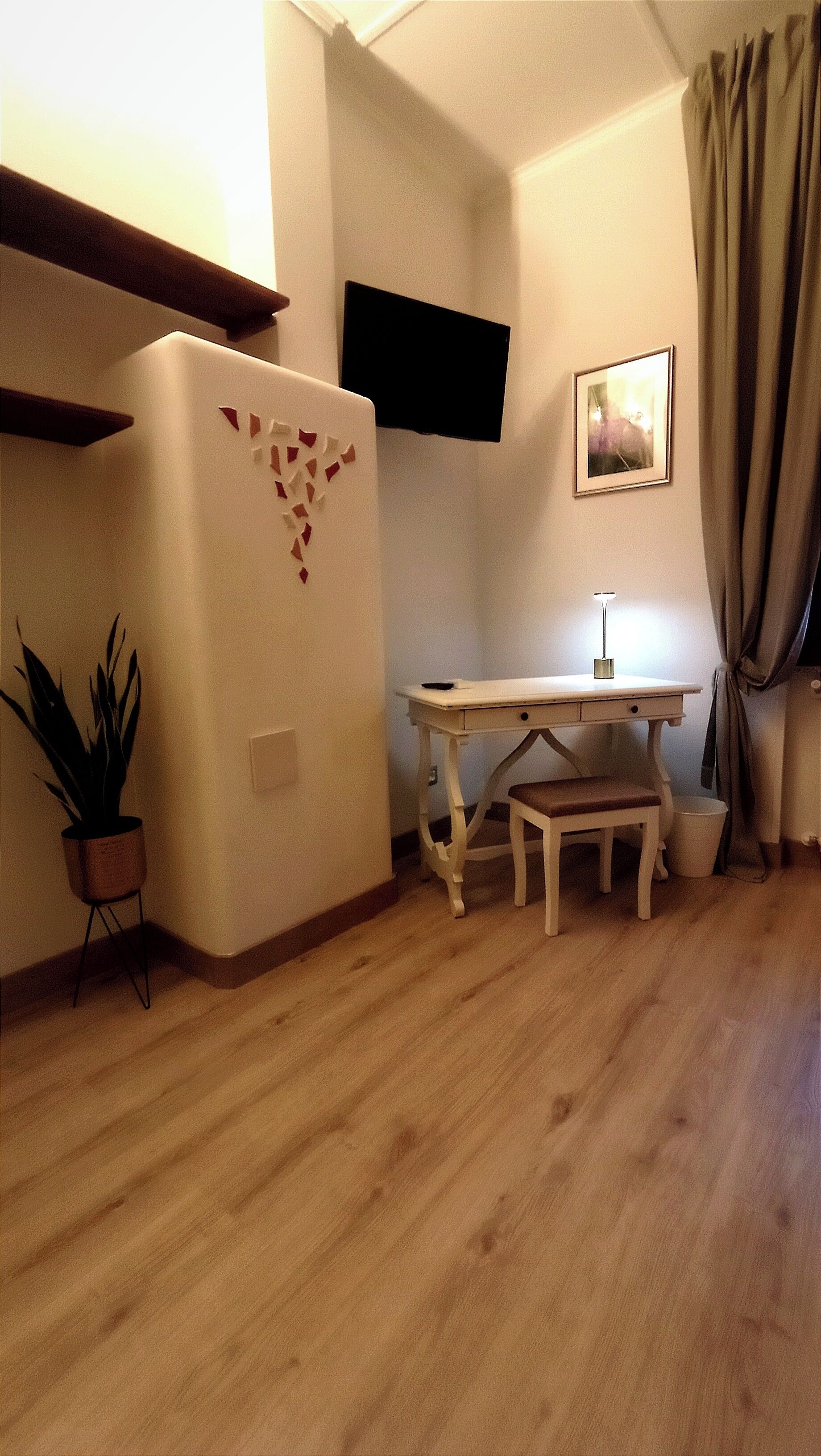 A small, cozy room with a wooden floor, a white desk with a chair, a small table, a potted plant, a white refrigerator, and a picture on the wall.