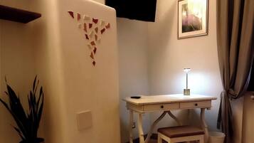 A small, cozy room with a wooden floor, a white desk with a chair, a small table, a potted plant, a white refrigerator, and a picture on the wall.