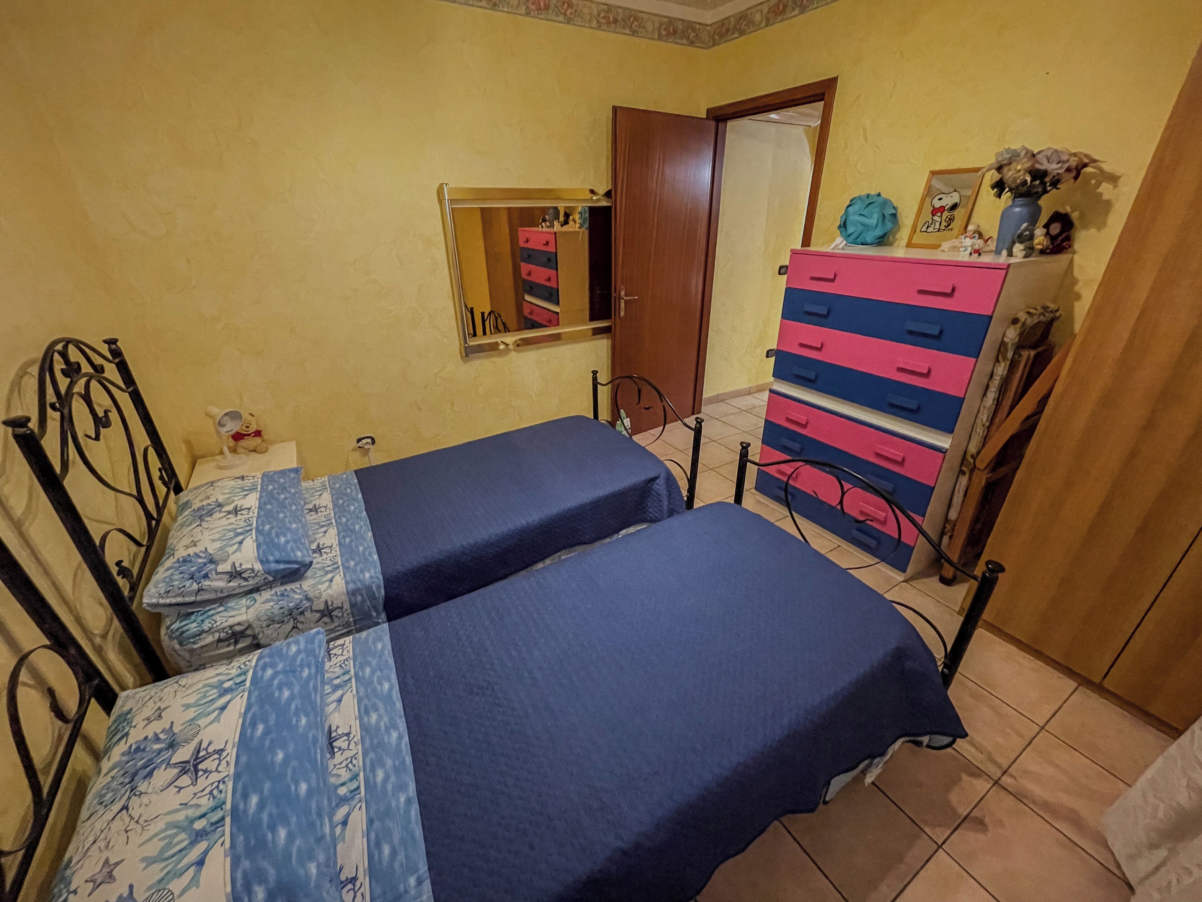 A bedroom with two single beds, a dresser, a mirror, and a door leading to another room.