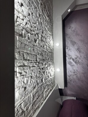 A room with a textured stone wall, a bed with a purple bedspread, and a modern light fixture.