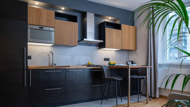 A modern kitchen with dark cabinetry, a stainless steel range hood, and a wooden countertop. There are two bar stools and a large potted plant.
