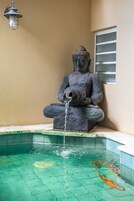 Una estatua de Buda con un desagüe de agua, un estanque de carpas y una ventana con persianas horizontales.