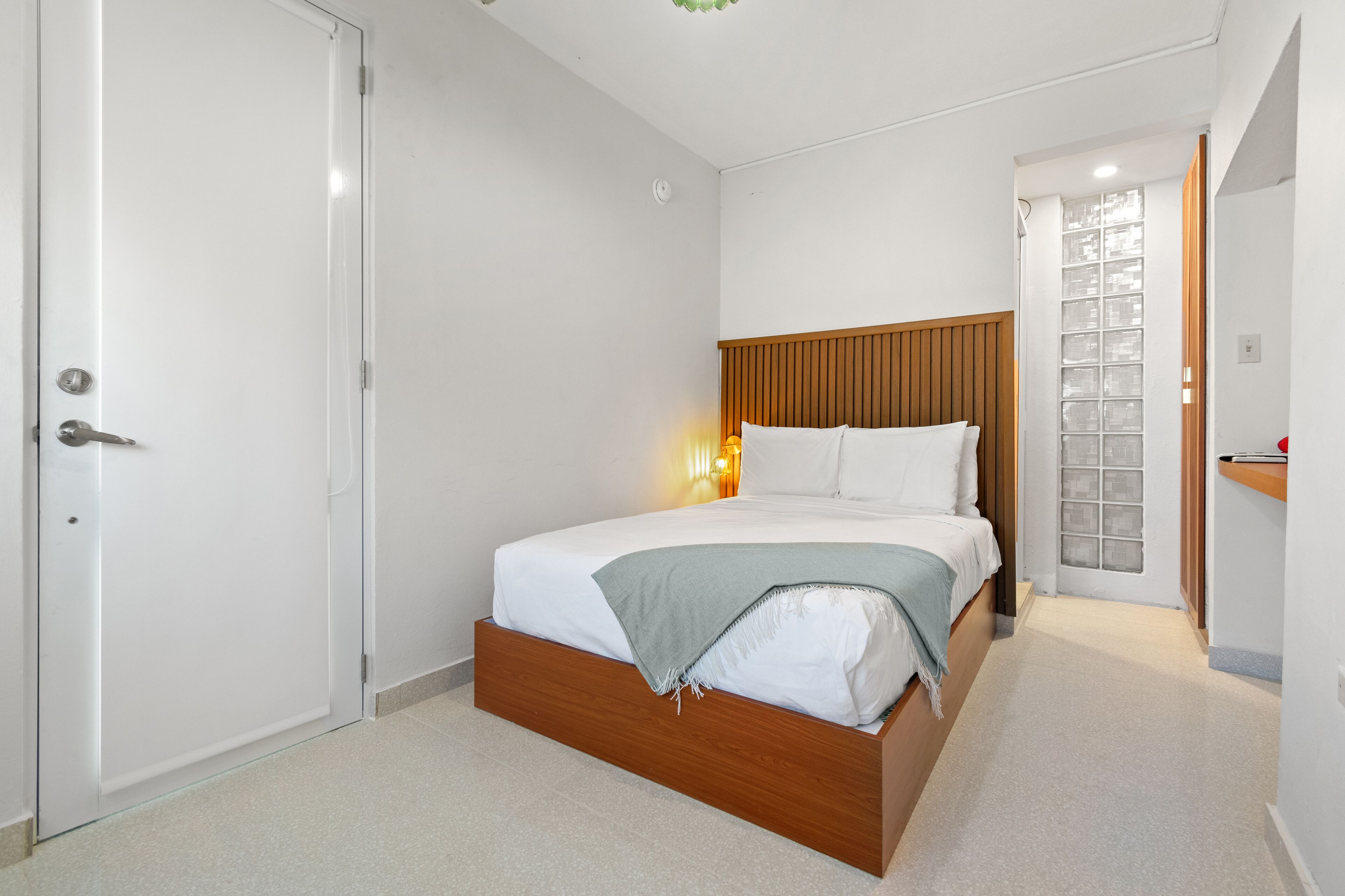 A bedroom with a wooden bed frame, white bedding, and a nightstand with a lamp.