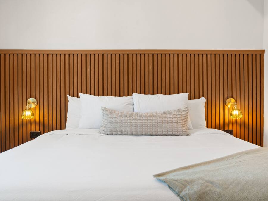 Una cama bien tendida con sábanas blancas y una manta color beige, con dos lámparas de noche de pantalla amarilla, frente a un cabecero de madera.