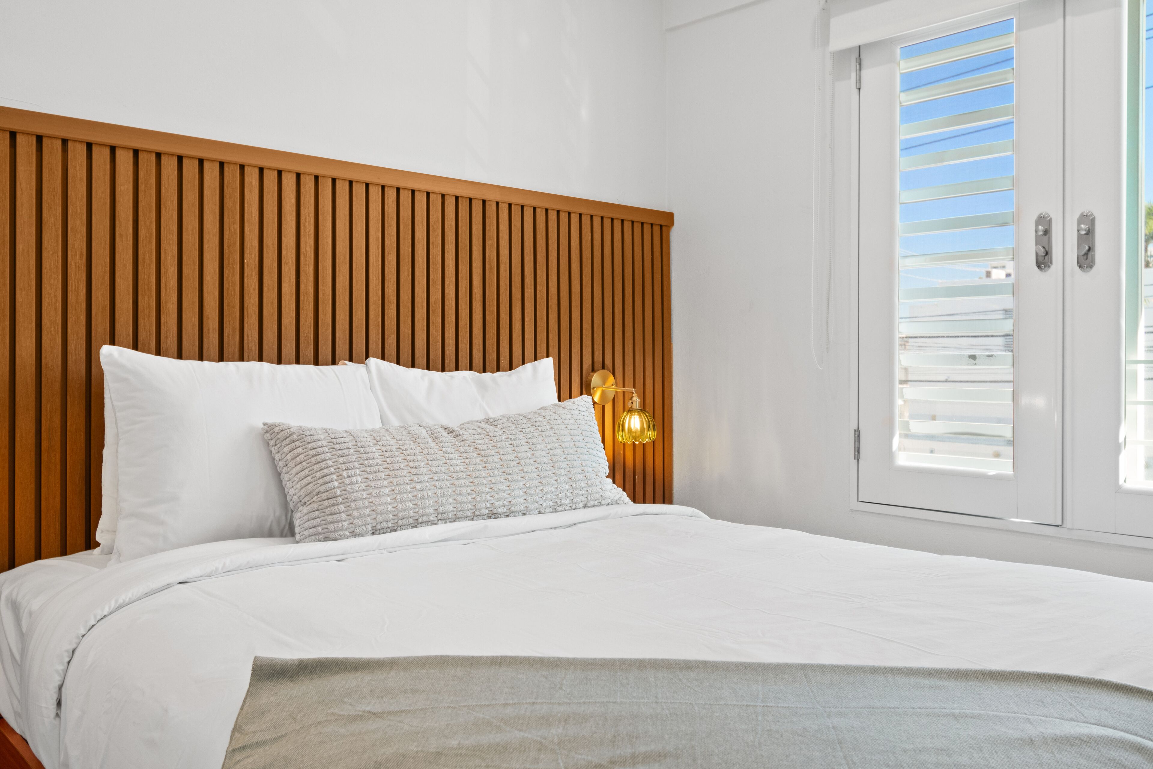 Un dormitorio con cabecera de madera, ropa de cama blanca, una lámpara de noche y una ventana con persianas.