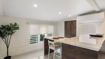 A modern kitchen with a white countertop, wooden cabinets, and a built-in stove. There is a window with blinds, a potted plant, and a white chair.