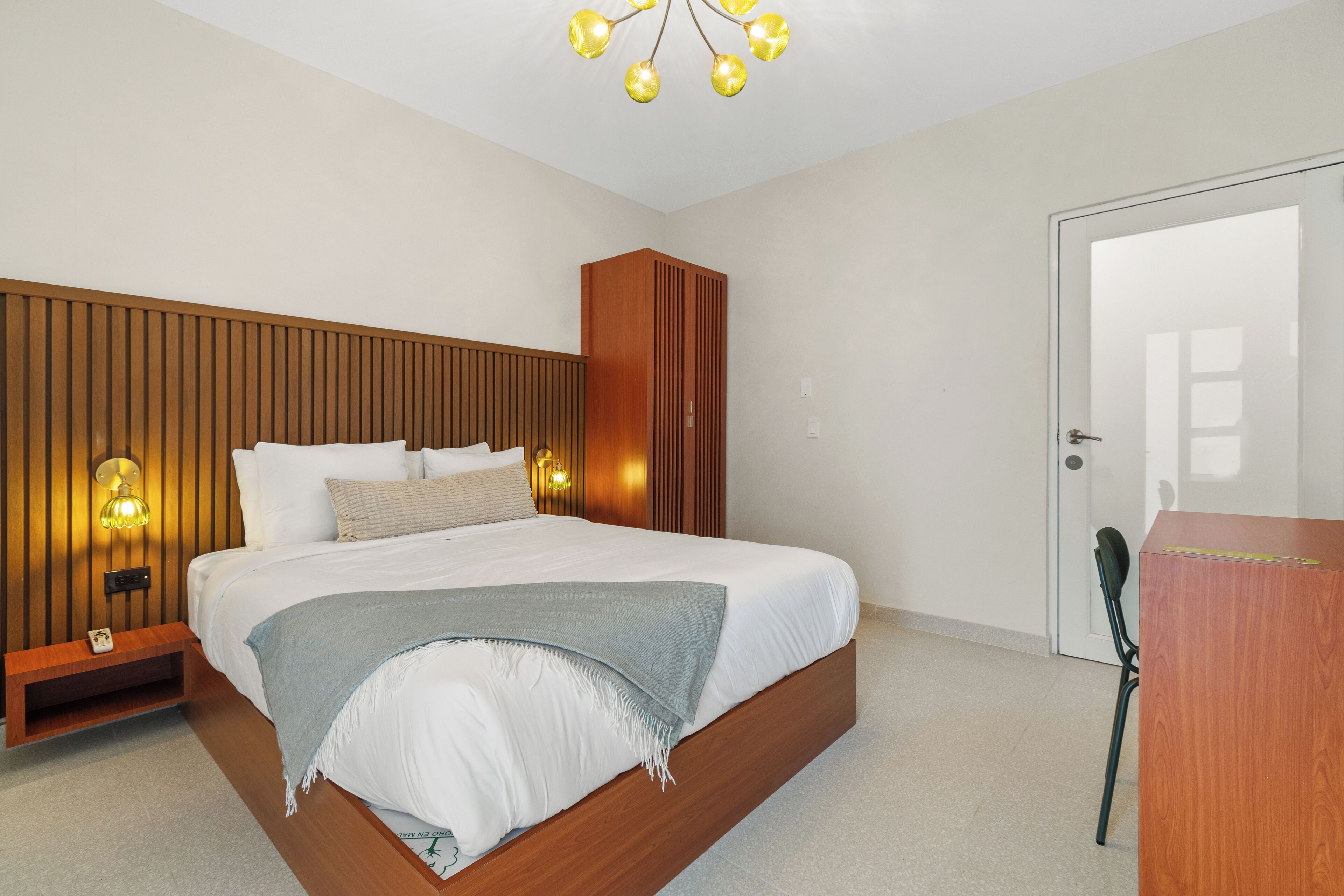 Ein Schlafzimmer mit einem hölzernen Kopfende, einem Bett mit weißer Bettwäsche, einer grauen Decke und einem Nachttisch mit einer Lampe.
