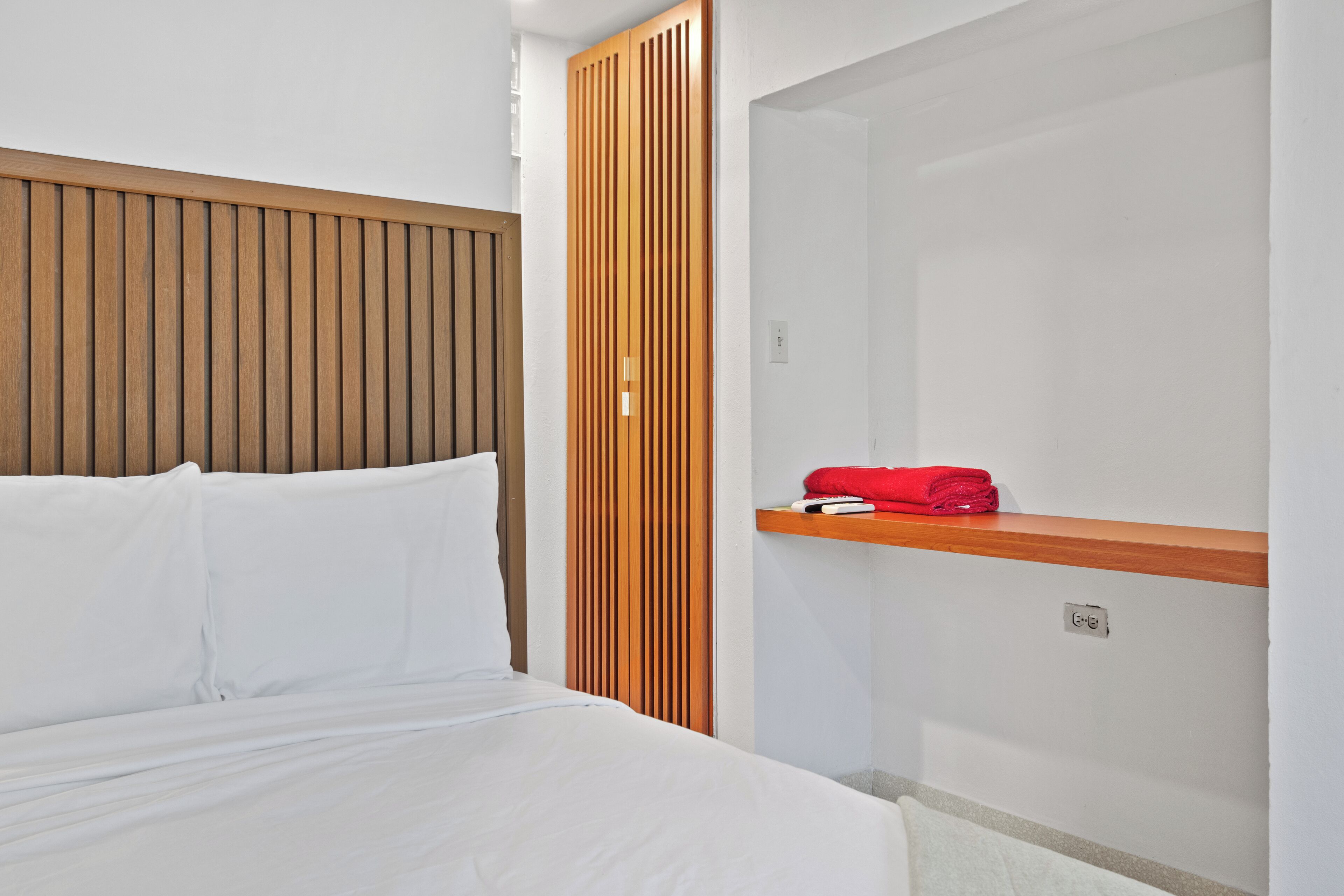 Habitación de hotel con cama, cabecero de madera, panel de pared de madera, repisa con toalla roja y pared blanca con interruptor de luz.