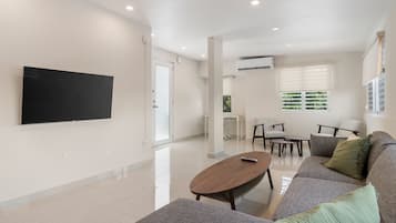 A modern living room with a flat-screen TV, a wooden coffee table, a grey sofa, and a dining area with chairs and a table.