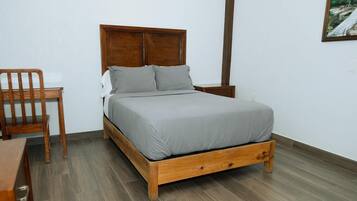 A single bed with a wooden frame and a grey mattress in a room with wooden flooring and a white wall.