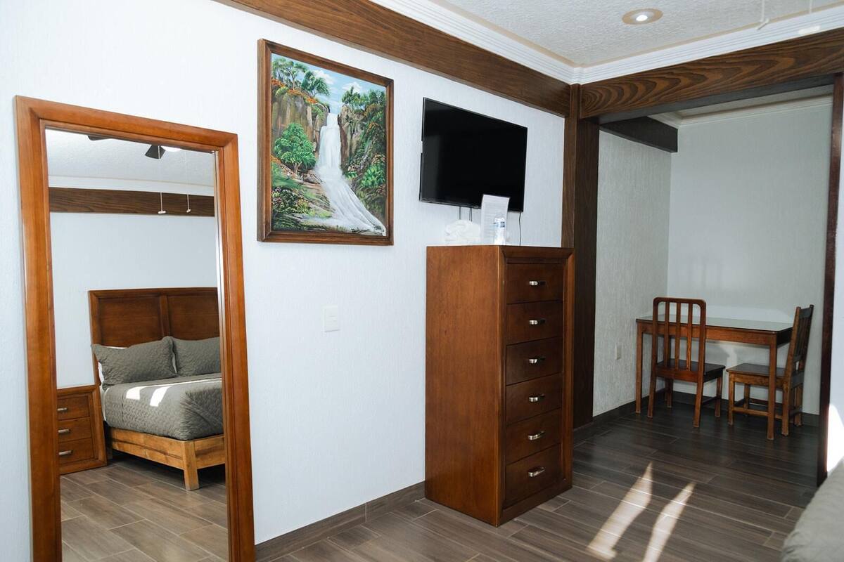 Un dormitorio con cama de madera, cómoda, televisor montado en la pared y una zona de comedor con mesa y sillas.