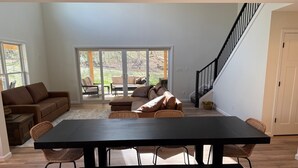 Amplia sala de estar con un gran comedor negro, múltiples opciones de asientos que incluyen un sofá y sillas, una escalera y un piso de vidrio corredero que da a un área exterior.
