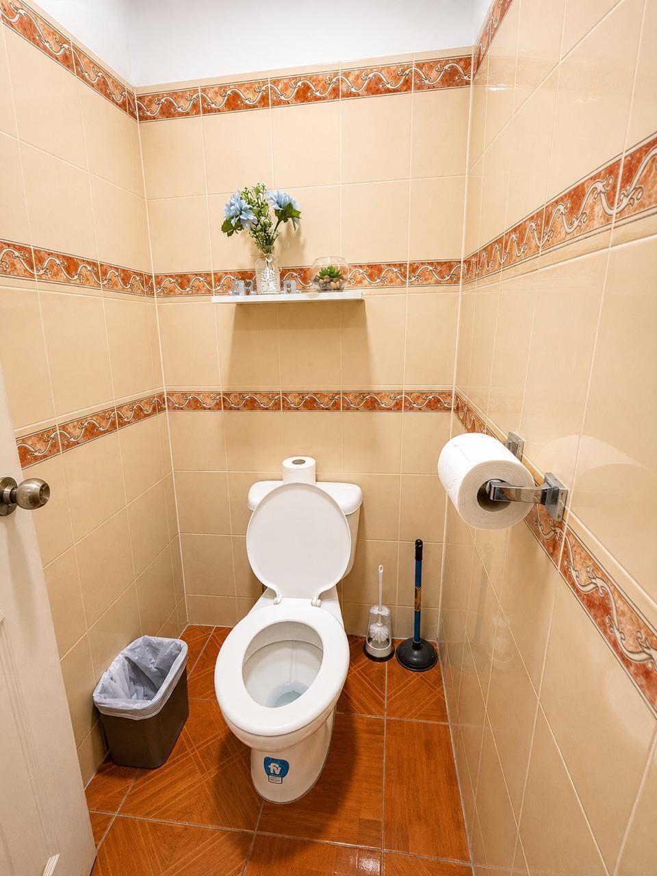 A small bathroom with a toilet, a trash bin, a toilet paper holder, and a small shelf with a plant.