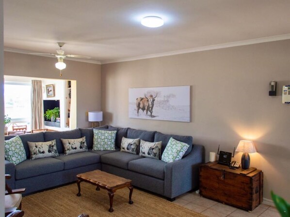 A living room with a grey sofa, patterned cushions, a wooden coffee table, a painting on the wall, and a lamp on a wooden side table. - Golfbeach Luxury Self Catering Apartment (Cape Town)