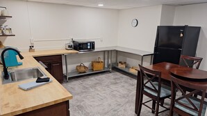 Een kleine keuken met een spoelbak, magnetron en zwarte koelkast. Er is een houten eettafel met stoelen en een plank met rieten manden.