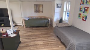 Een kamer met een houten vloer, een bed met een grijze quilt, een bureau met een laptop en een kleine plant, een schilderij aan de muur en een raam met uitzicht.