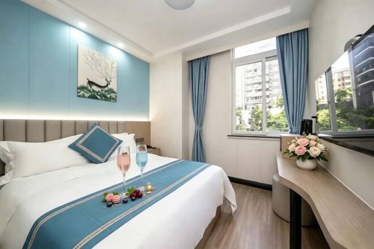 Une chambre d’hôtel avec un lit, une table de chevet ornée d’un vase de fleurs, une fenêtre avec des rideaux et une vue sur la ville.