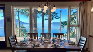A dining area with a table set for four, large windows offering a view of a lake and trees, and a chandelier above.