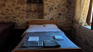 A stone-walled bedroom with a wooden bed, a shelf above the bed, and a window with curtains. - Fani's house (Platanos)