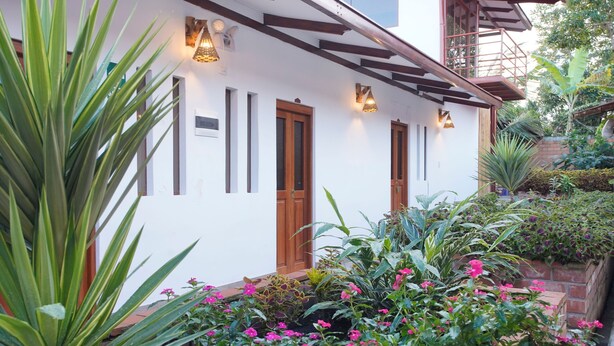 A white building with wooden doors and windows, surrounded by lush greenery and flowering plants. - Muyuterra (Moyobamba)