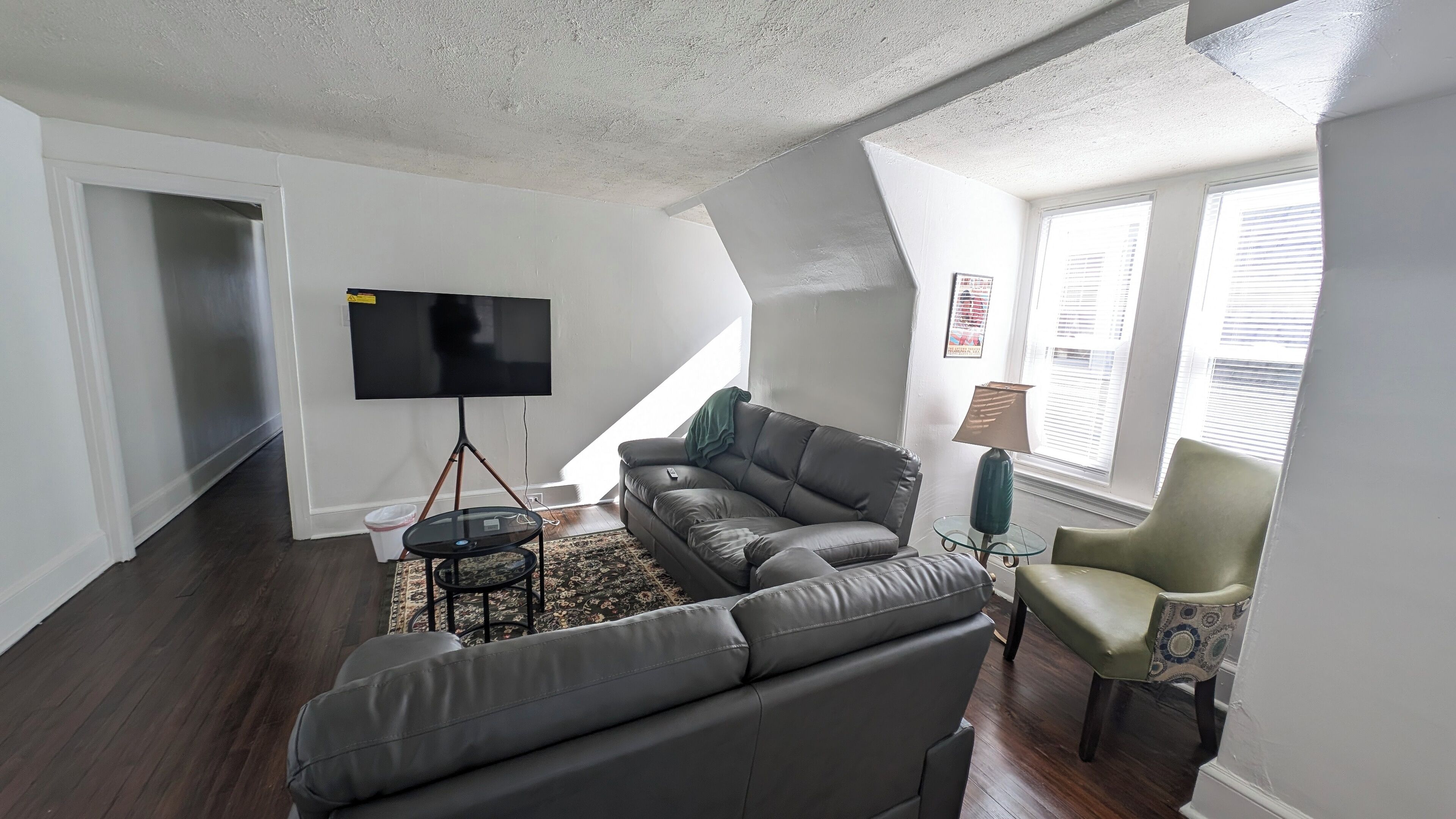 Una sala de estar con un televisor de pantalla plana grande sobre un soporte tipo trípode, un sofá de cuero oscuro, una mesa de centro de vidrio, un tapete estampado, un sillón verde y una lámpara sobre una mesita.