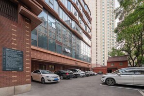 Un parking rempli de voitures, un bâtiment doté de grandes fenêtres et, au fond, un grand immeuble d’habitation.