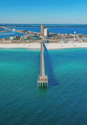 An aerial view of a pier extending into turquoise waters, with a beach and buildings in the background.