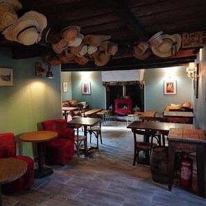 Um restaurante acolhedor com mesas e cadeiras de madeira, uma lareira e chapéus pendurados no teto.
