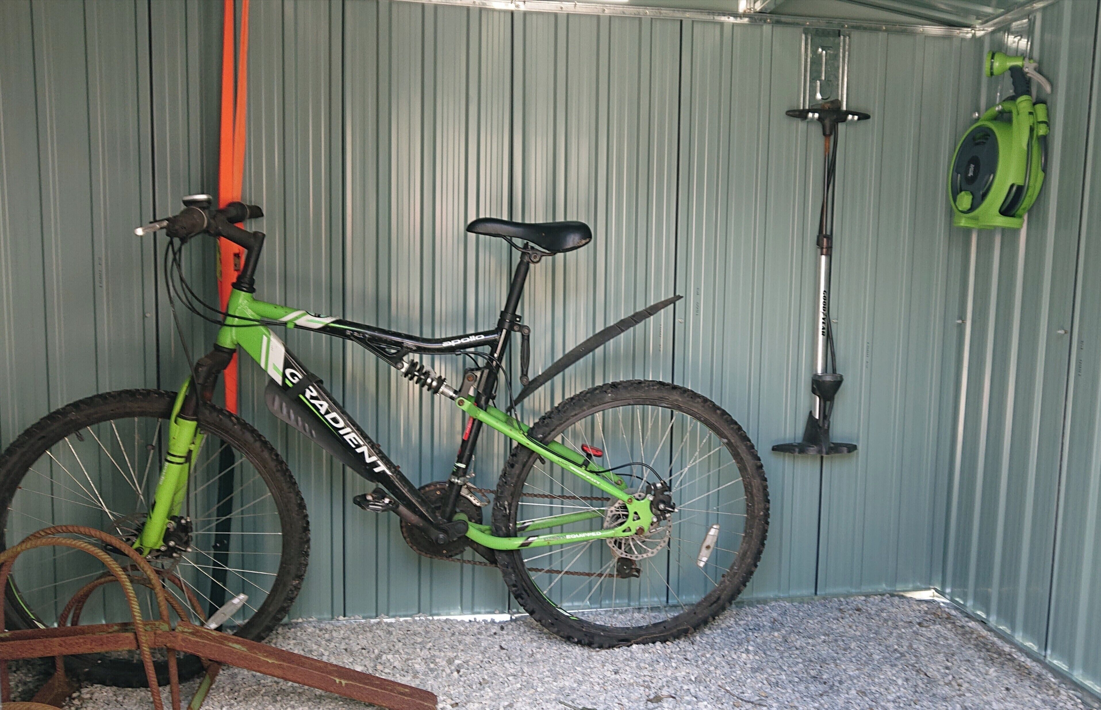 A bicycle parked against a corrugated metal wall with a green and black frame, a water pump, and a green garden tool hanging on the wall.