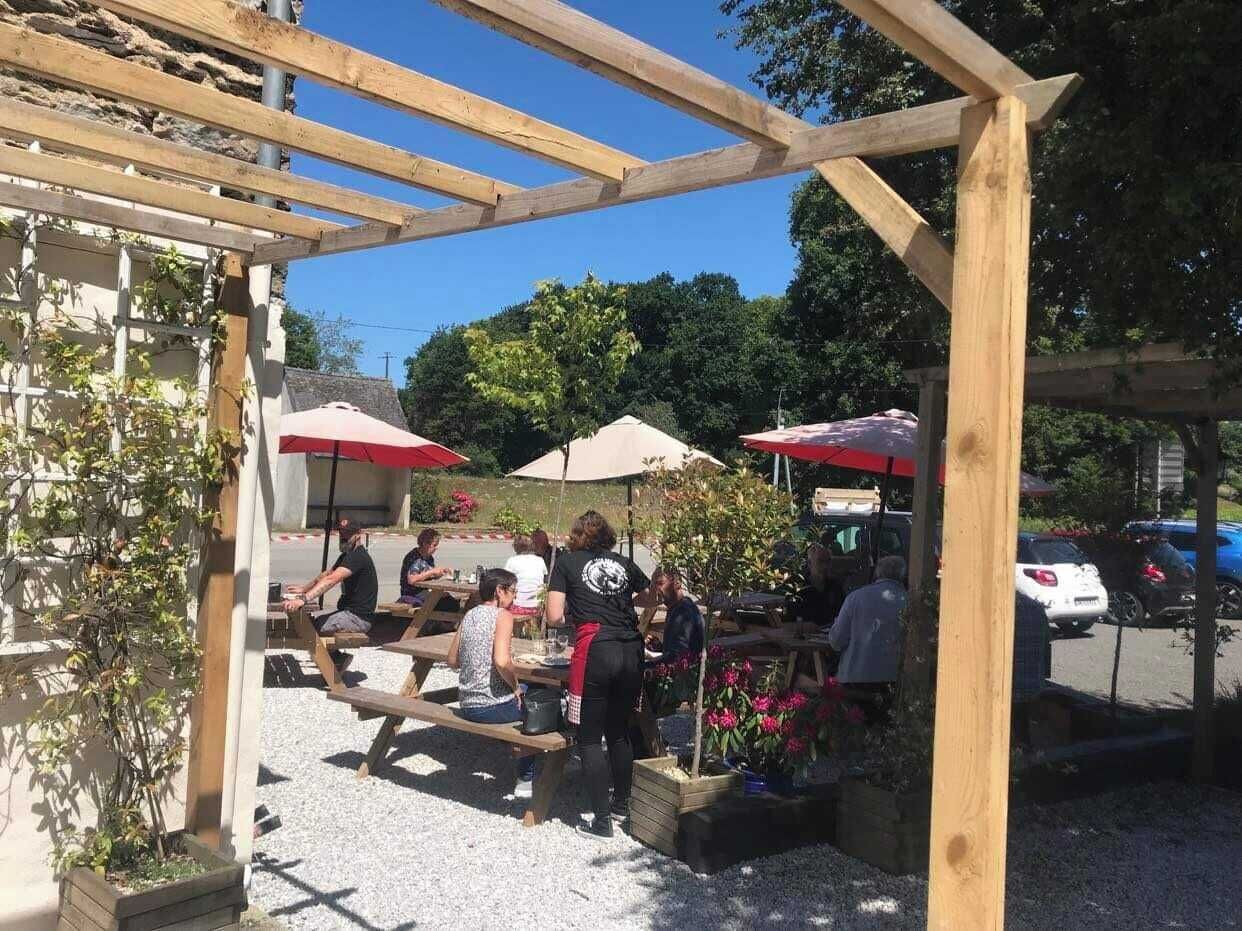 Un espace extérieur aménagé avec des tables en bois et des parasols rouges, entouré de verdure et bordé d’un parking.