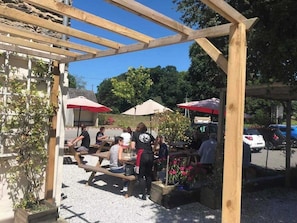 An outdoor seating area with wooden tables and red umbrellas, surrounded by greenery and a parking lot.