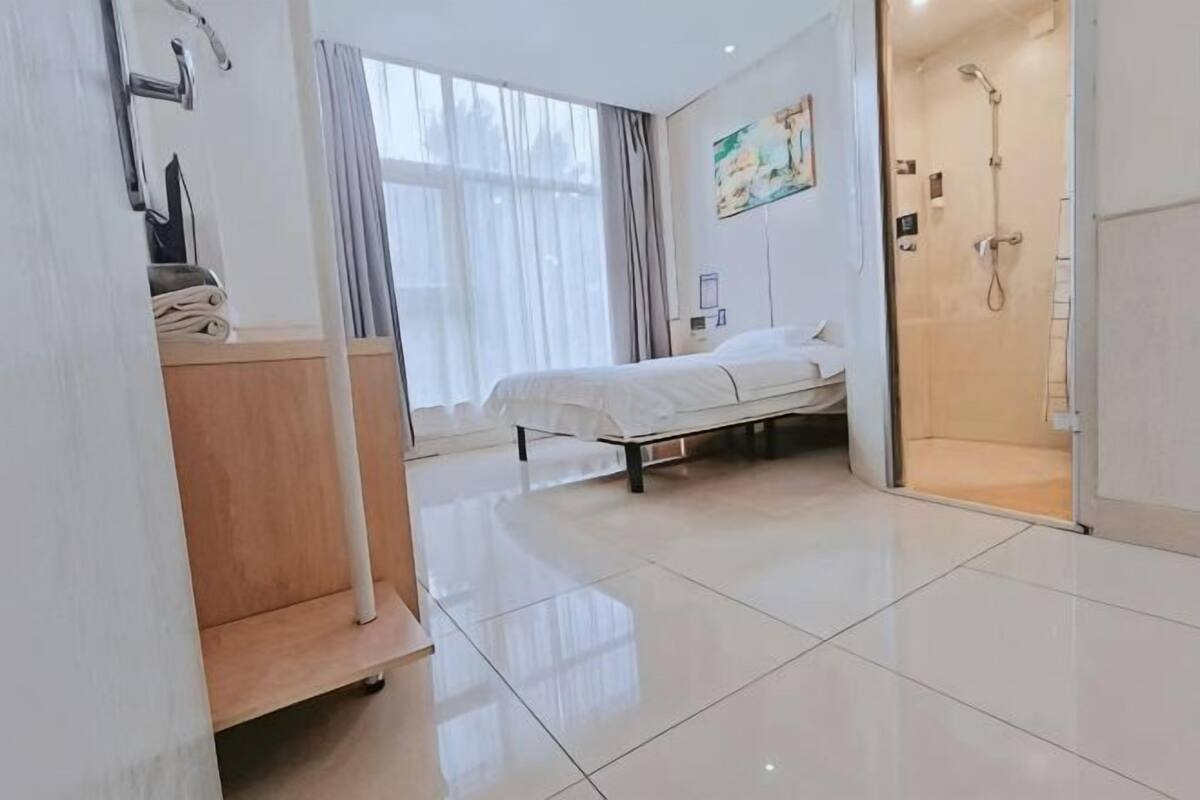 A hotel room with a bed, a shower, a wooden cabinet, and a large window with curtains.