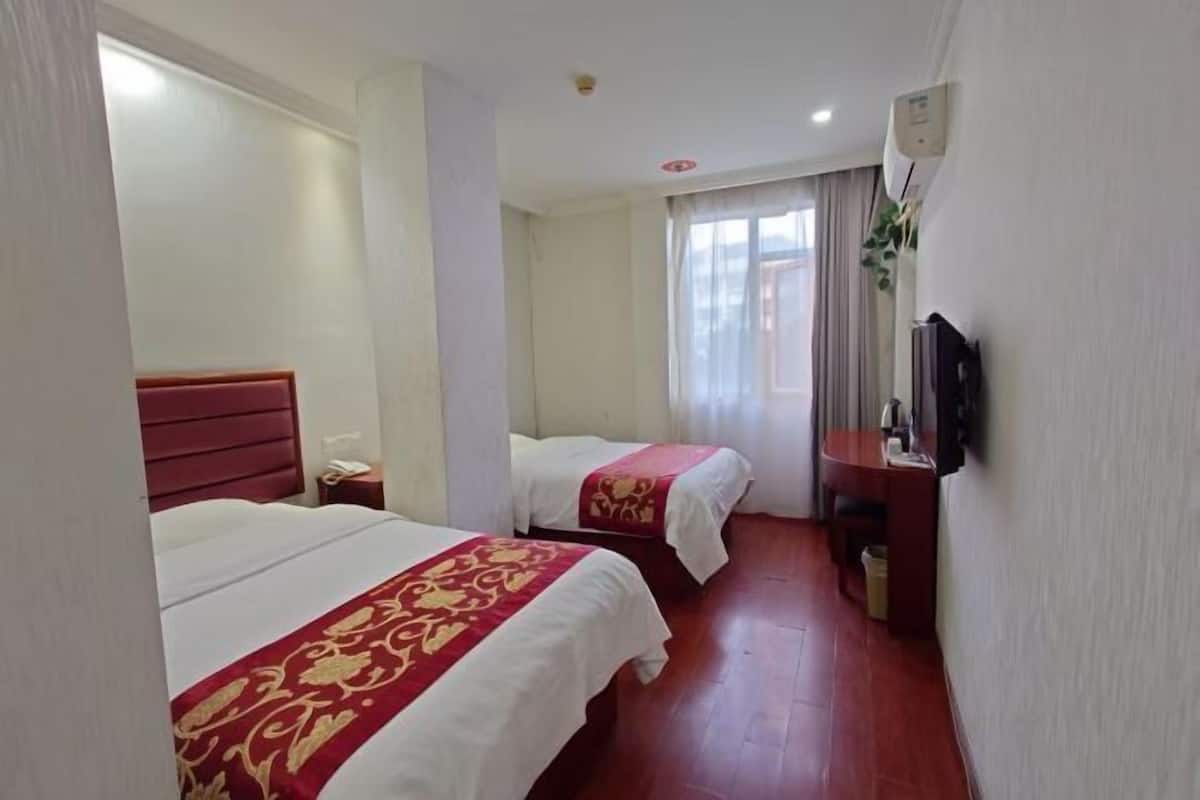 A hotel room with two beds, a wooden headboard, a bedside table with a phone, a TV mounted on the wall, and a window with curtains.