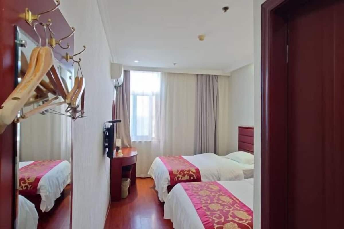 A hotel room with two single beds, a wooden wardrobe, a desk with a chair, and a large window with curtains.