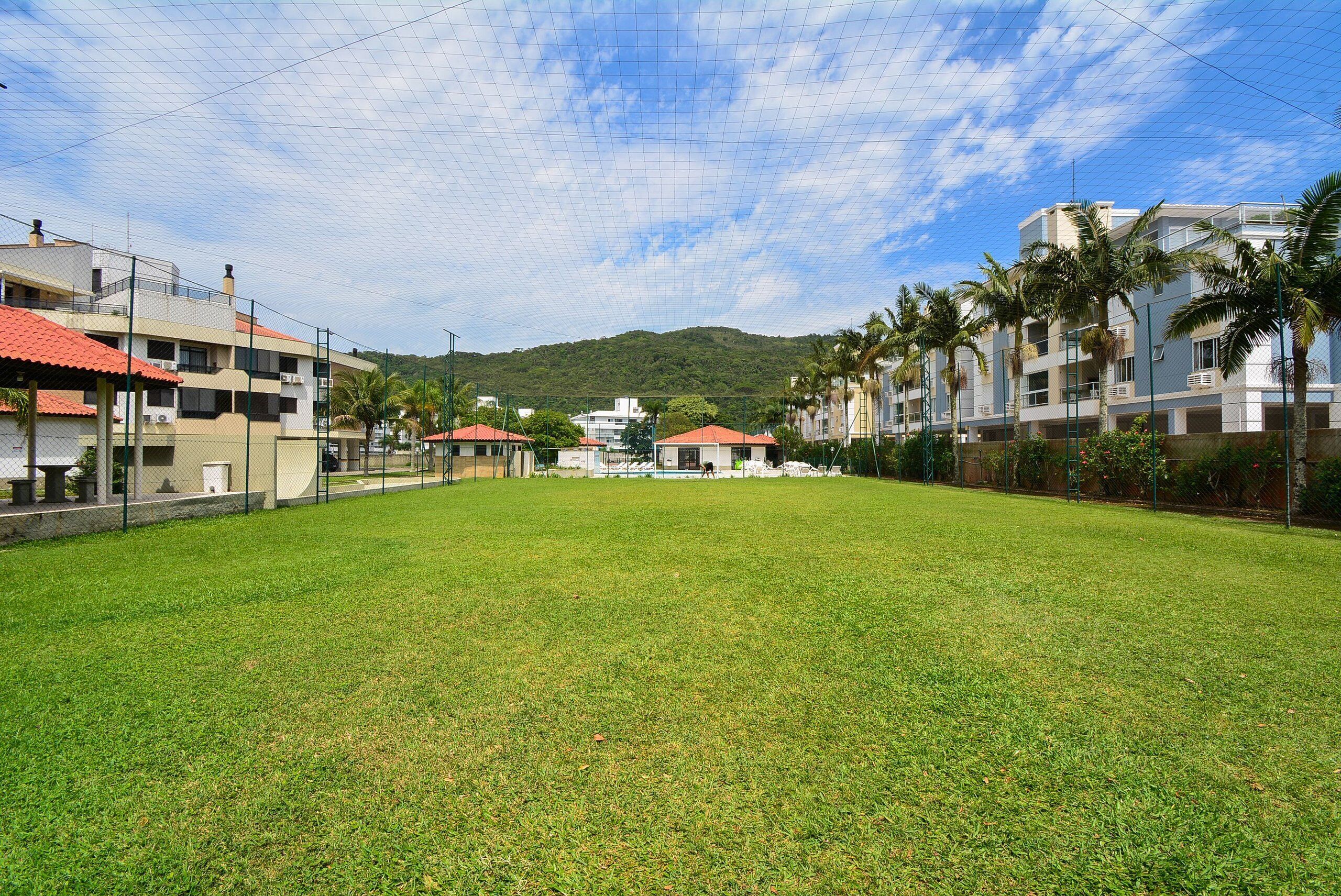 A grassy area with buildings in the background, including a white building with a red roof and a multi-story building with balconies and palm trees.