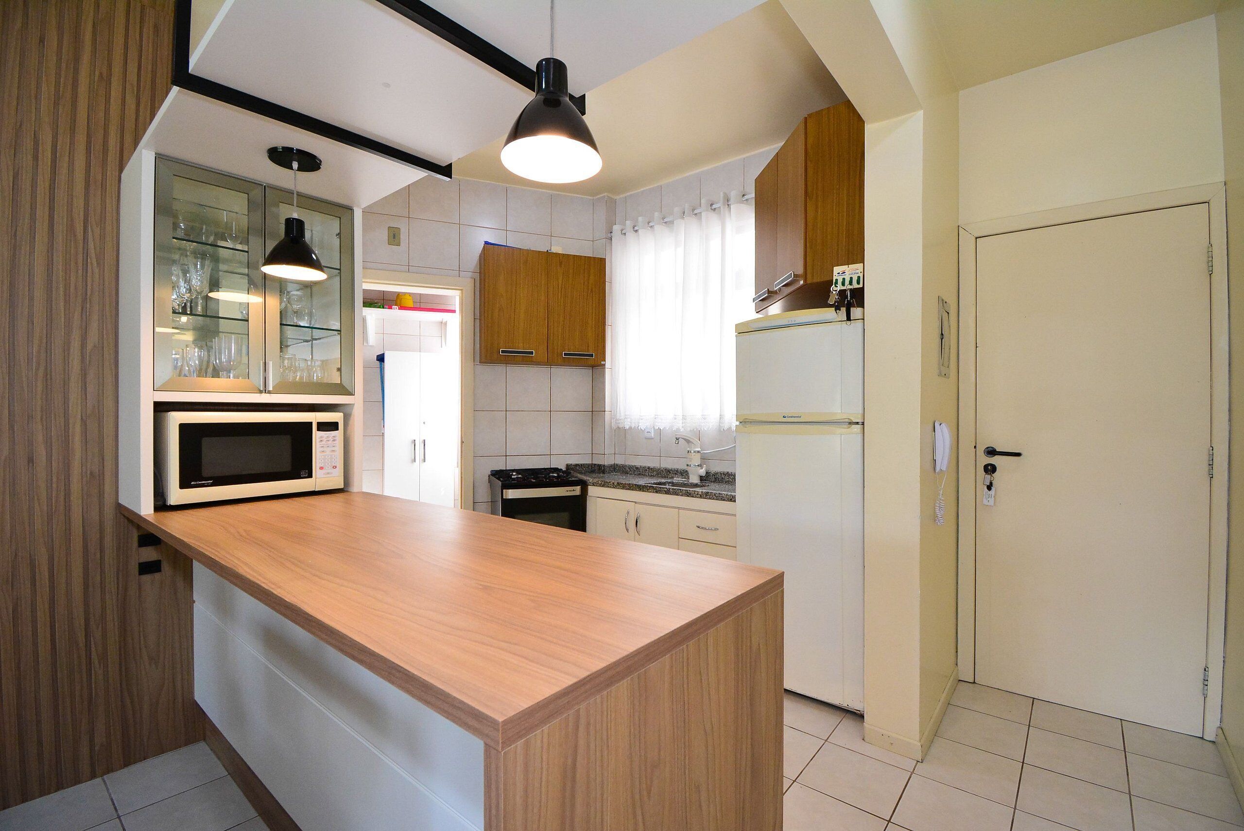 Una cocina compacta con encimera de madera, microondas, estufa y refrigerador. Se aprecia un armario con puertas de vidrio y un colgante de luz.