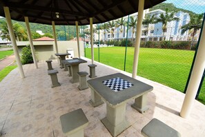 Un patio al aire libre con una mesa de ajedrez y bancos, rodeado por una cerca verde y palmeras.