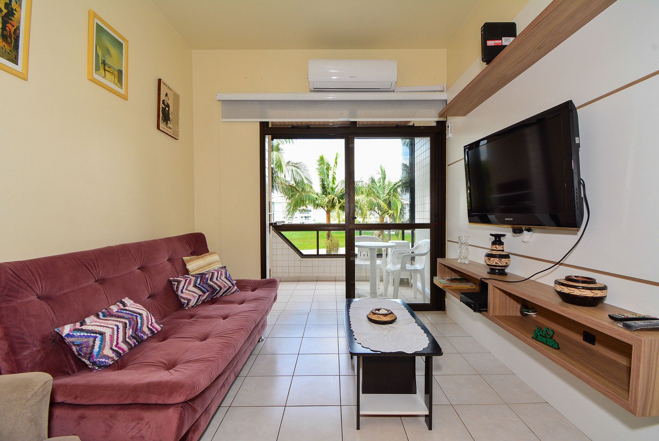 A living room with a red sofa, a coffee table, a TV, and a balcony with a view of palm trees.