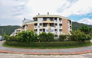 Un edificio de varios pisos con balcones, rodeado de abundante vegetación y un telón de fondo montañoso.