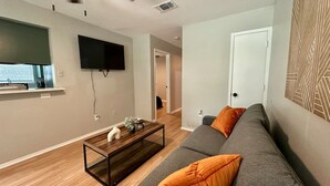 Un salon moderne avec un canapé gris, des coussins orange, une table basse en bois, un téléviseur à écran plat et un ventilateur de plafond.