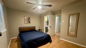 Une chambre à coucher avec un lit, un ventilateur de plafond, un tableau encadré au mur et une porte donnant sur une autre pièce.