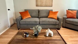 Un salon avec un canapé gris, des coussins orange, une table basse agrémentée d’une plante et d’un vase, et une œuvre d’art géométrique accrochée au mur.