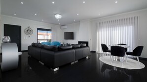 Ein modernes Wohnzimmer mit einem schwarzen Sofa, einem runden Tisch mit Stühlen, einem flachbildfernseher an der Wand und einem großen Fenster mit leichtem Vorhang.