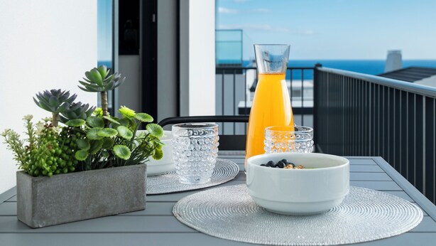 Ein Tisch mit einem Blumentopf, einem Saftglas und einer Schüssel Essen auf einem Balkon mit Meerblick.