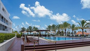 A pool area with lounge chairs and umbrellas, surrounded by palm trees and a modern building. - Solar Tambaú - LikeHome Hospedagens (Joao Pessoa)