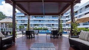 A covered outdoor seating area with tables and chairs, overlooking a swimming pool and a modern building with balconies. - Solar Tambaú - LikeHome Hospedagens (Joao Pessoa)