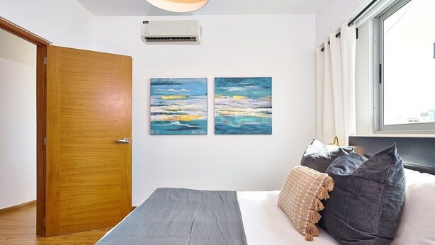 A bedroom with a bed, two wall-mounted paintings, a window with curtains, and a wooden door. - Torre Armonia 24 (Santo Domingo)