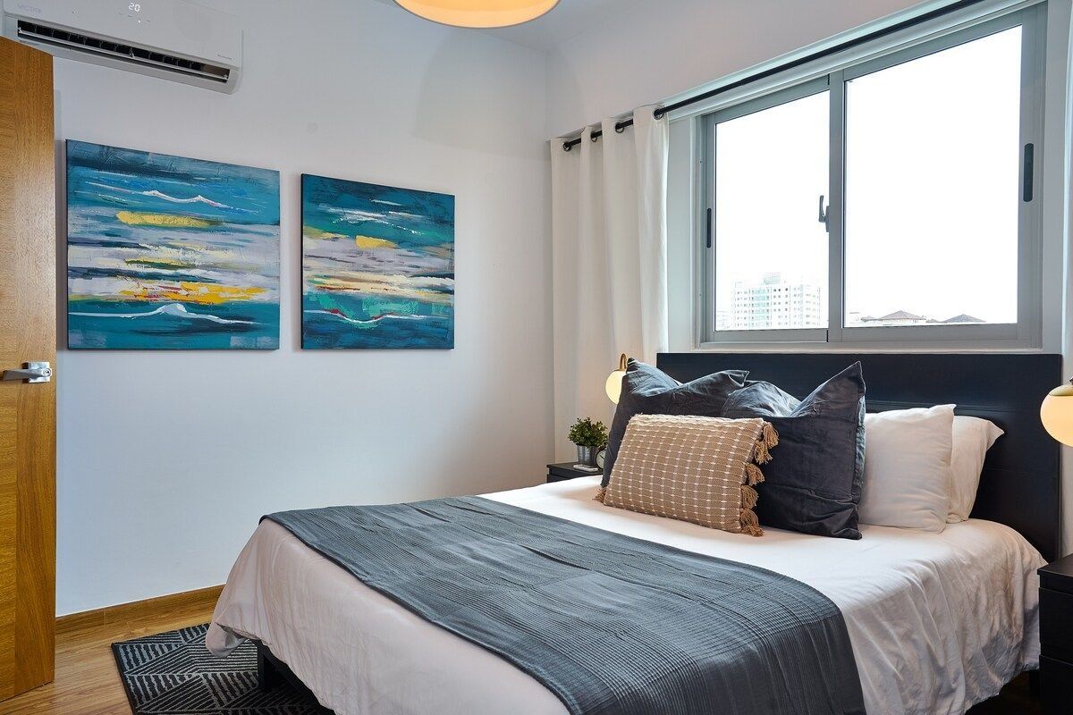 A bedroom with a bed, two paintings, a window, and a nightstand.