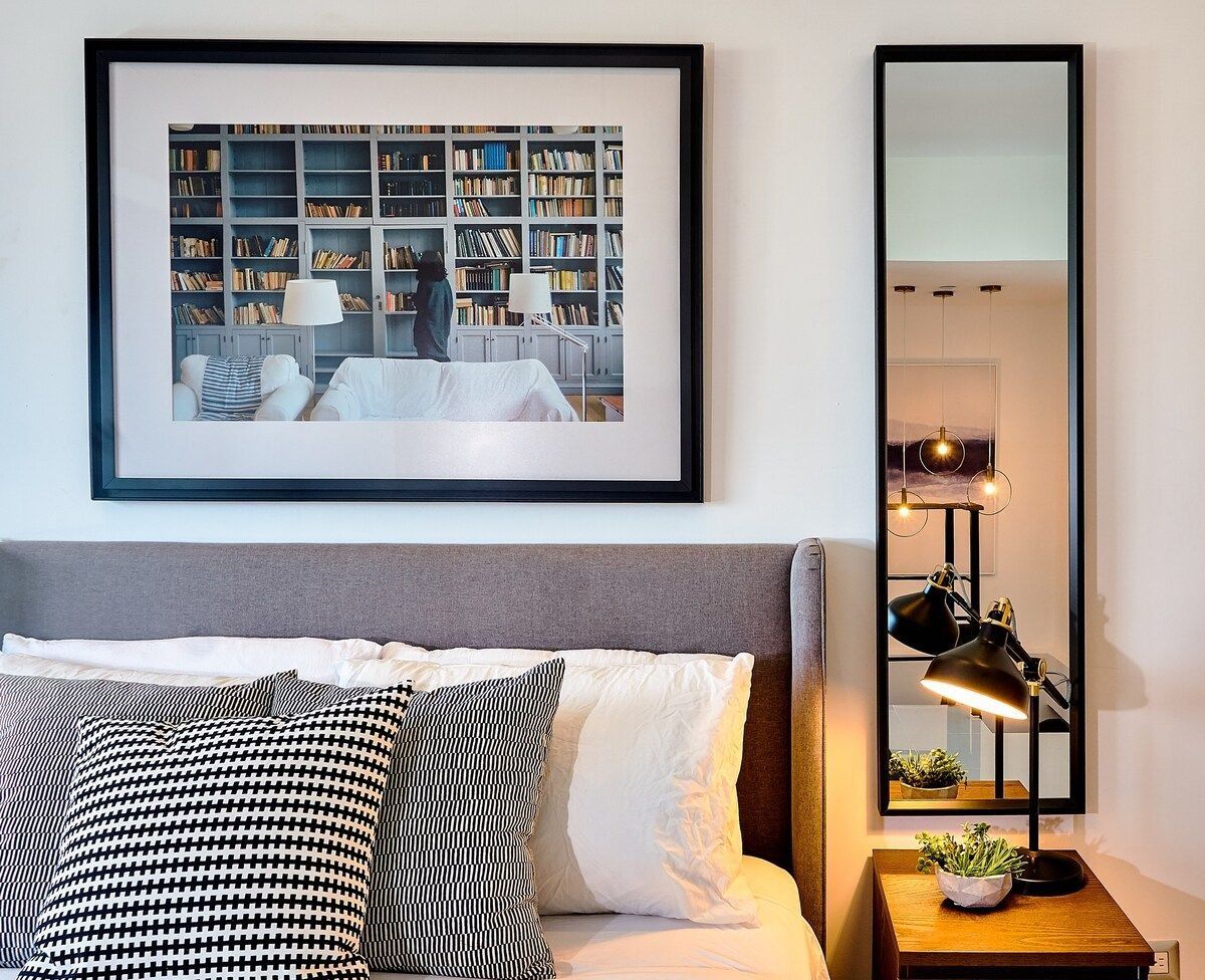 Ein ordentlich eingerichtetes Schlafzimmer mit einem Bett, Kissen, einem Nachttisch mit Lampe, einem gerahmten Bild an der Wand und einem Bücherregal.