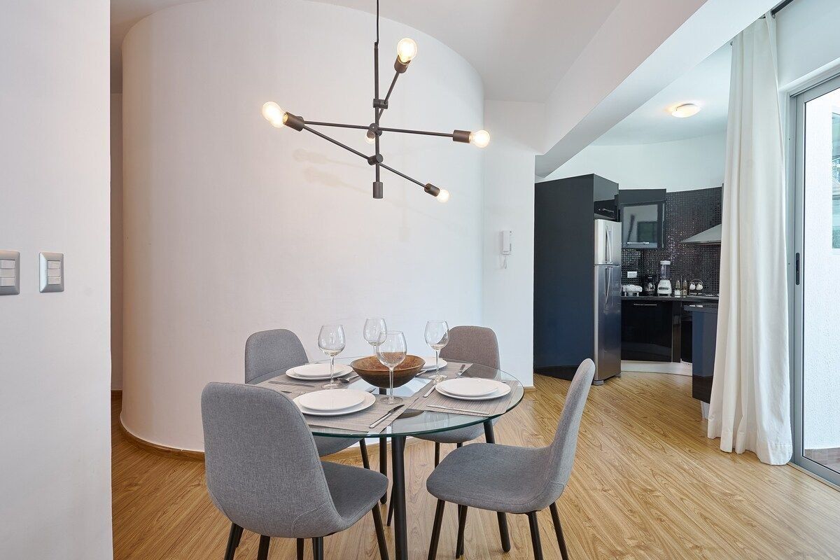 Un espace repas moderne avec une table ronde en verre pour quatre personnes, des chaises grises à revêtement en tissu et un lustre contemporain.